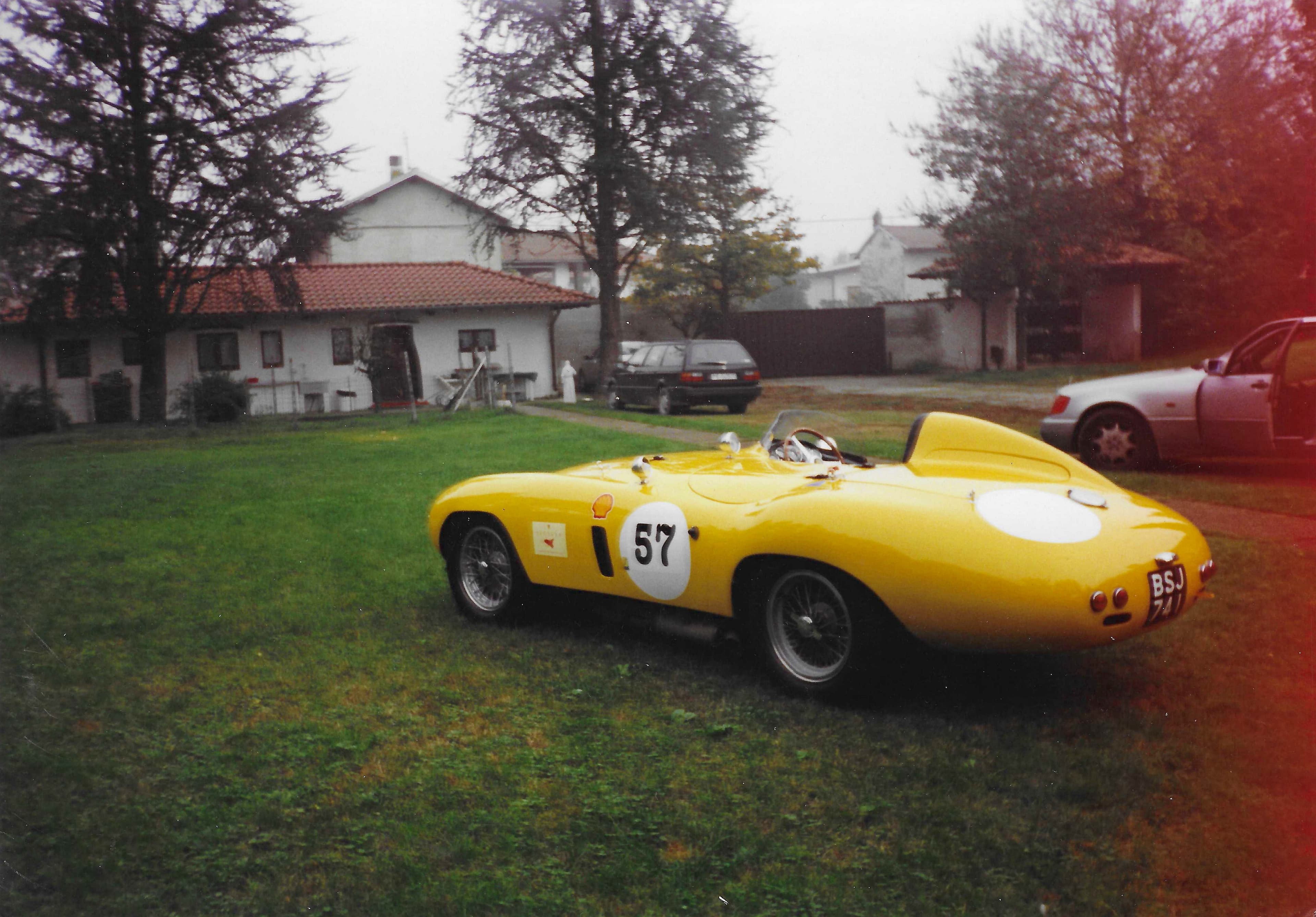 Bright yellow vintage race car with number 57 parked on grass before a house.