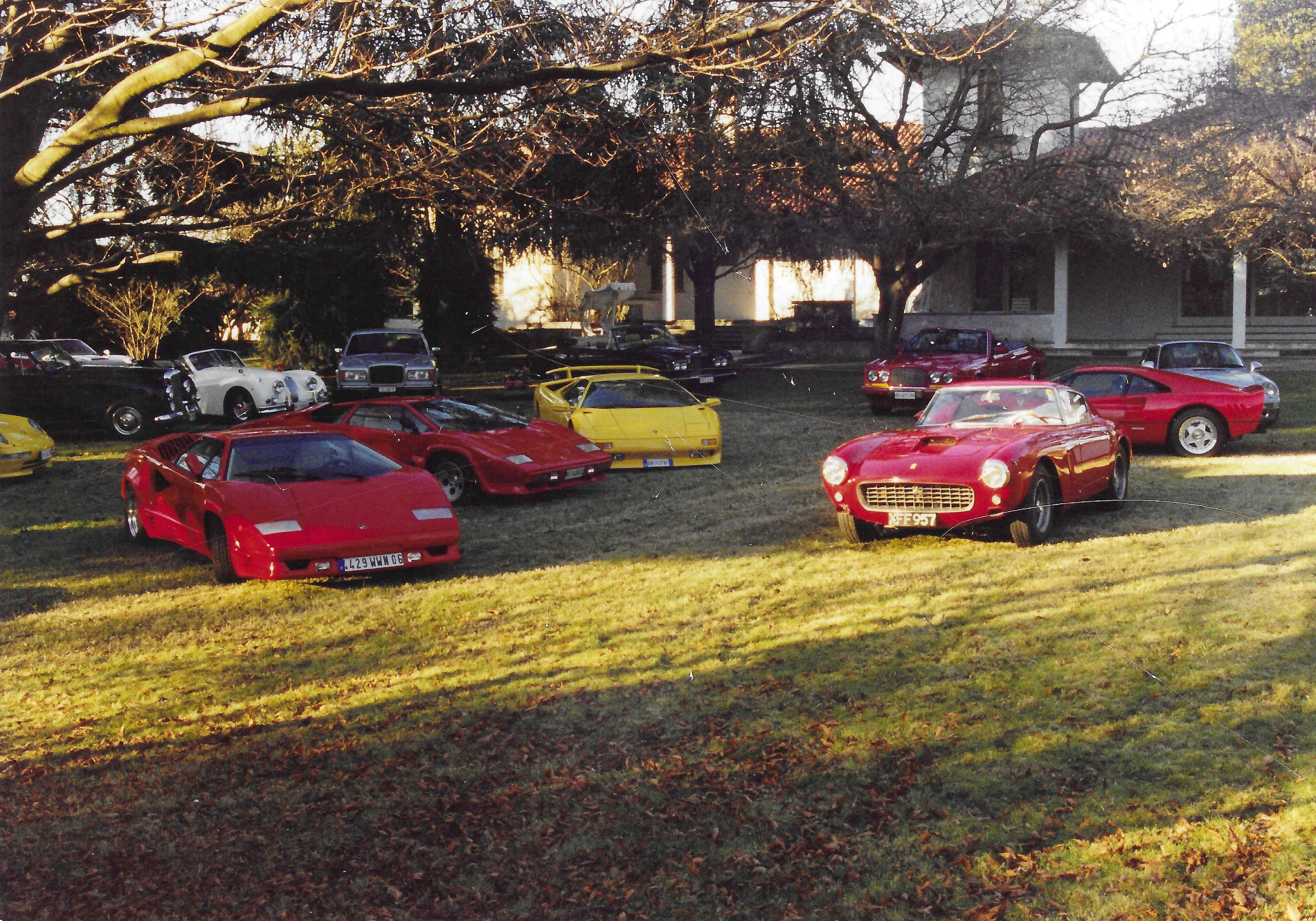 Luxury sports cars, including Lamborghinis and a Ferrari, parked on a sunlit grassy estate lawn.